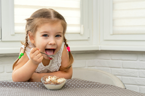 4 years old girl in a white blouse eagerly eats ice-cream from dumplings at a table in the kitchen and is happy