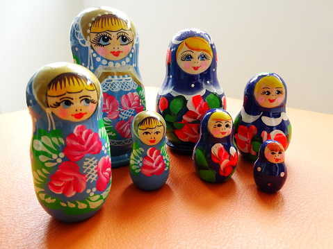 The photo shows seven Russian nesting dolls manufactured and painted by hand. The dolls characterize the generations of two different families. Vibrant, strong colors contrast with the delicacy of this souvenir. They are on an orange colored surface with a naturally lit white background.