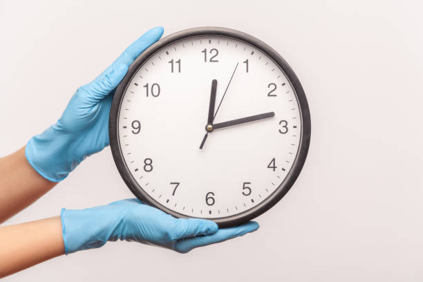 perfil de visión lateral primer plano de la mano humana en guantes quirúrgicos azules que sostienen el reloj analógico. - clock time alarm clock urgency fotografías e imágenes de stock