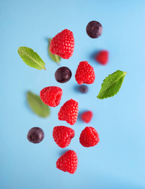 fliegende beeren auf blauem grund. fallende himbeerfrüchte und minze - falling fruit berry fruit raspberry stock-fotos und bilder