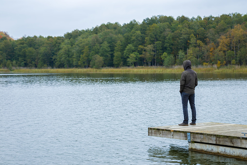 One young man standing alone on edge of wooden footbridge and staring at lake. Hooded guy. Thinking about life. Peaceful atmosphere in nature. Enjoying fresh air in cold cloudy day. Back view.