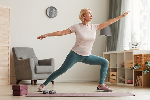 Mature woman standing on exercise mat and doing yoga in the living room at home
