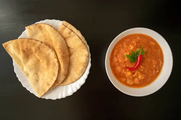 Photo of peeled fava beans with chili in white bowl and pita bread on dark wooden background
