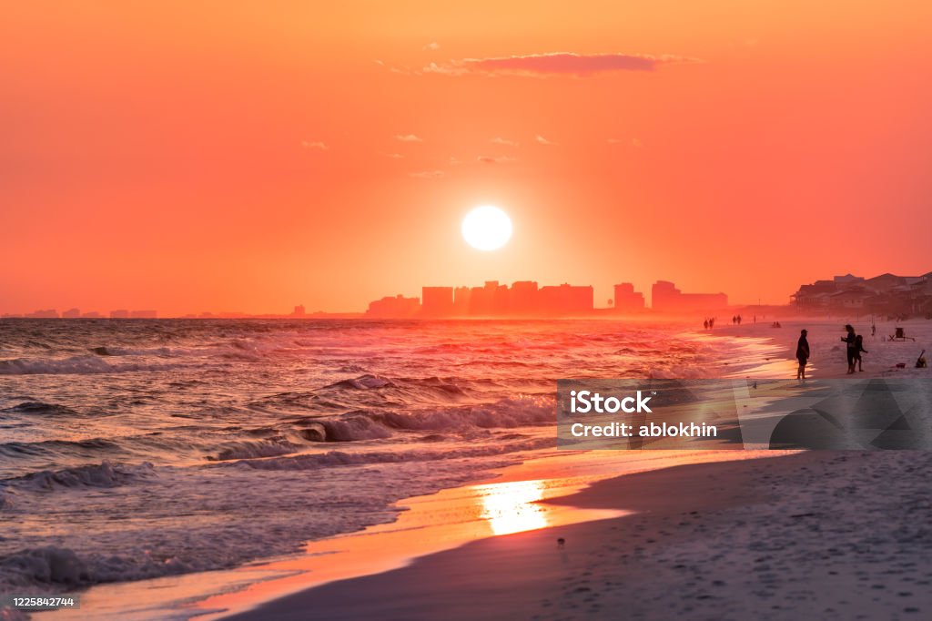 Couple walking on shore with dramatic orange red pink sunset in Santa Rosa Beach, Florida with Pensacola coastline coast cityscape skyline in panhandle with ocean gulf mexico waves Pensacola Stock Photo