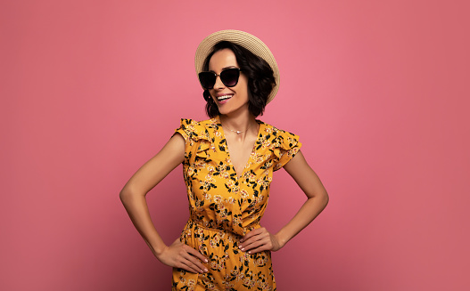 Sunglasses.. Close-up photo of a cheerful young girl in a floral dress, sunglasses, and straw hat, who is standing with her hands on her hips and smiling.