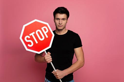 No more bad habits. Close-up photo of a serious young man in a black t-shirt, who is looking in the camera while holding a stop sign.