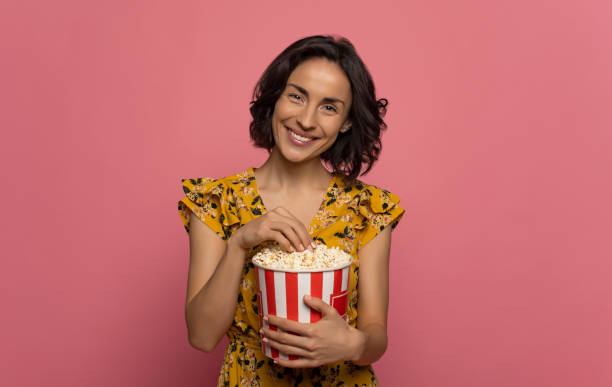 Eating habits. Close-up photo of a cheerful young lady, who is looking in the camera with a big smile, while eating popcorn. stock photo