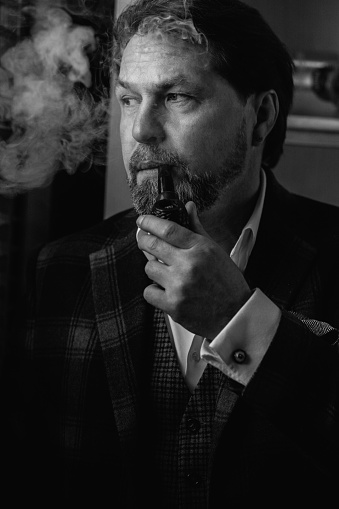White and black portrait of well-dressed mature bearded caucasian man, small business owner, in expensive tailored suit relaxing with tobacco pipe