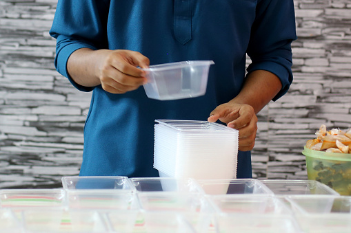A focus scene on Muslim teenage boy is preparing plastic containers to fill in 'nasi kerabu' (blue pea steamed rice with side vegetable dishes accompanied with fried chicken and chilli sauce) at home. He is assisting his mother whom a home-based chef and accomodate orders during Ramadan season coincide with partial lockdown in Malaysia.