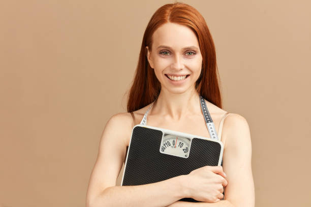 Beautiful young woman holding scale and too big trousers as she lost weight Young woman with unhealthy look looking happy and cheerful, holding scale in hands, isolated over beige background too thin stock pictures, royalty-free photos & images
