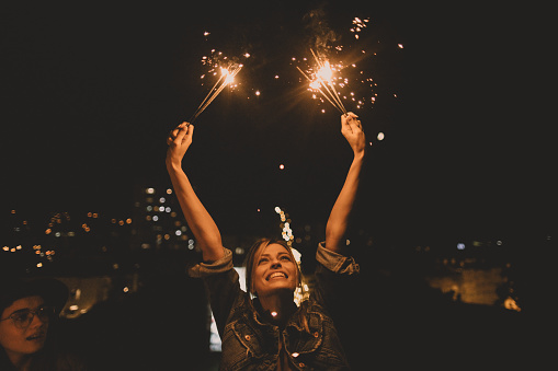 Photo of a happy woman holding sparklers while enjoying the rooftop party with her girlfriends; celebrating her youth and femininity.