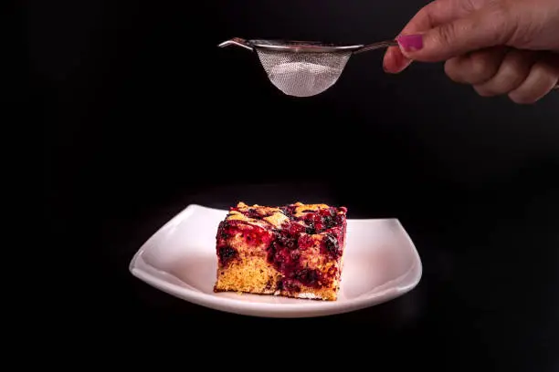 woman's hand sprinkling white powder sugar on homemade cake with forestfruit served on a plate with a spoon