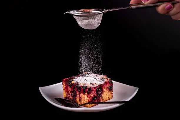 woman's hand sprinkling white powder sugar on homemade cake with forestfruit served on a plate with a spoon