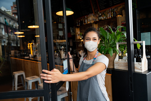 Happy business owner opening the door at a cafe wearing a facemask to avoid the spread of coronavirus â reopening after COVID-19 concepts