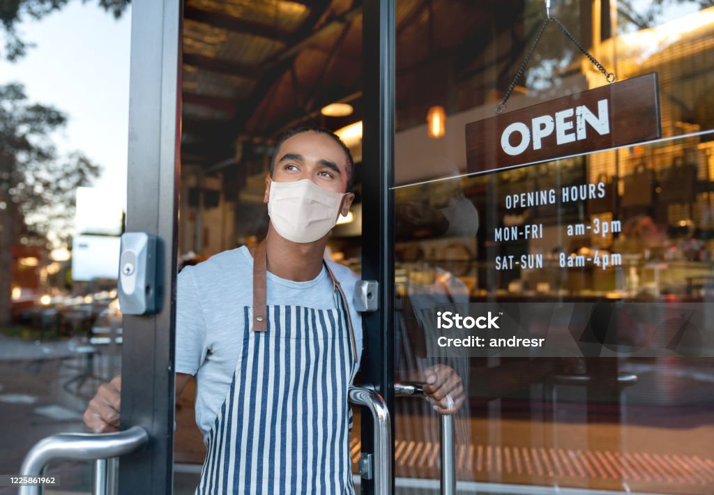 Business owner opening the door at a cafe wearing a facemask Business owner opening the door at a cafe wearing a facemask to avoid the spread of coronavirus â reopening after COVID-19 concepts Business Stock Photo