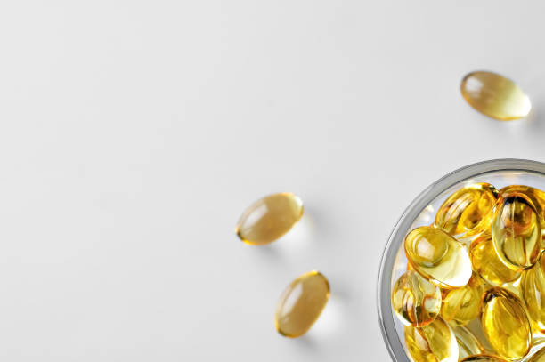 Capsules of fish oil (omega-3) in a glass bowl and on a white stschl nearby. Flat lay with copy space, white background. stock photo
