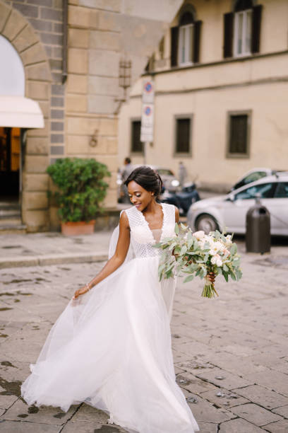 matrimonio a firenze, italia. sposa afroamericana in un abito bianco con un lungo velo e un mazzo di fiori tra le mani. - abito da sposa foto e immagini stock