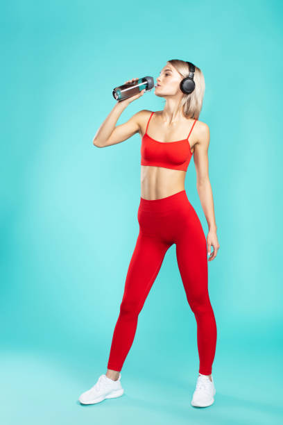 So thirsty. Young and sporty blonde woman in headphones and red sportswear drinking water while standing against blue background. Full length So thirsty. Young and sporty blonde woman in headphones and red sportswear drinking water while standing against blue background. Music and sport. Fitness. Workout. Studio shot blonde female bodybuilders stock pictures, royalty-free photos & images