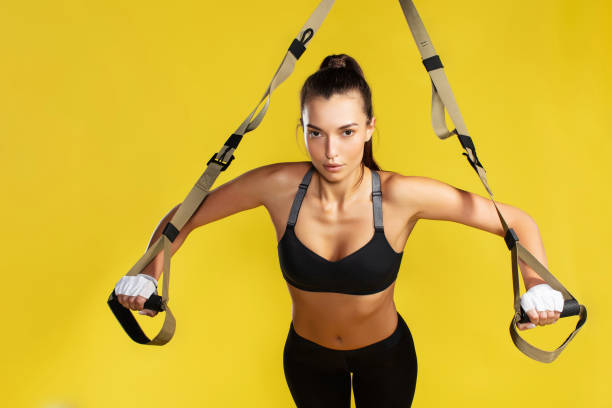 Training arms. Strong young woman in sportswear doing push ups with trx fitness straps against yellow background. She is looking at camera. Studio shot Training arms. Strong young woman in sportswear doing push ups with trx fitness straps against yellow background. She is looking at camera. Studio shot. Sport, training, trx, fitness. Workout suspension training stock pictures, royalty-free photos & images