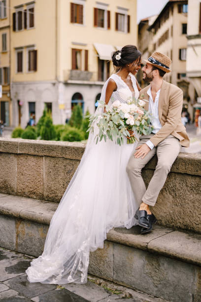 인종 간 결혼식 부부. 피렌체에서 결혼식, 이탈리아. 긴 베일과 꽃다발을 입은 흰색 드레스를 입은 아프리카계 미국인 신부와 모래 색 재킷과 밀짚 모자를 쓴 백인 신랑. - 웨딩 드레스를 뉴스 사진 이미지