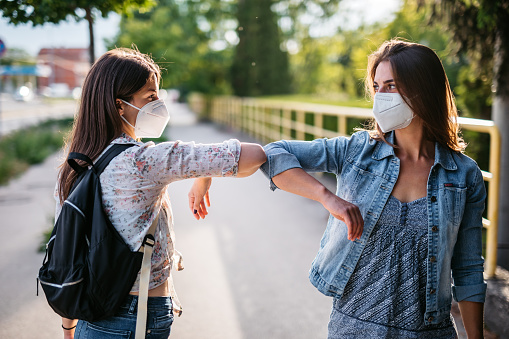 Two young female Caucasian friends greeting alternatively during pandemic and wearing n-95 face masks.