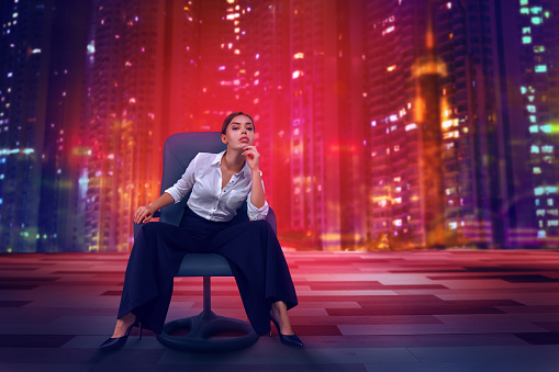 Beautiful businesswoman sitting on armchair in modern office with futuristic night city in background. Young woman sit on office chair
