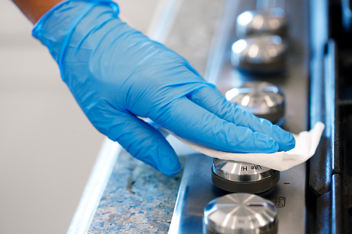 A man wearing a blue protective glove cleans a stove top knob with a disinfectant wipe.