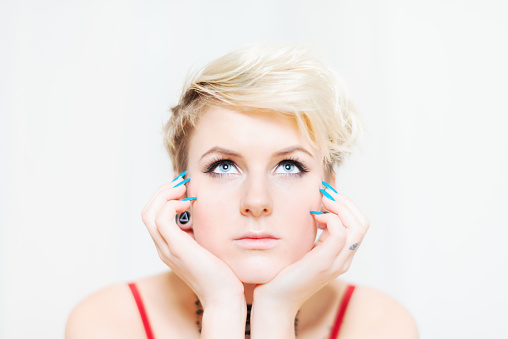 Young woman with bright blue eyes, blonde short hair and turquoise blue fingernails looking up, thinking with her head in her hands. Studio Shot, White Background.