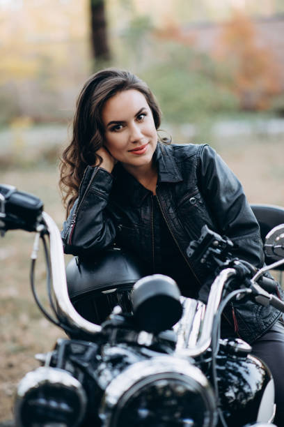 joven hermosa motociclista con una chaqueta de cuero negro en una motocicleta con un casco en las manos. el concepto de velocidad y libertad. enfoque selectivo suave. - motorcycle women helmet sensuality fotografías e imágenes de stock