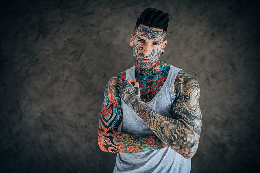 One man, modern man with whole body in covered in tattoos, posing by the gray wall.