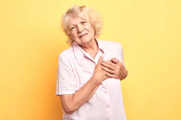Gray haired old beautifullady in white shirt suffers from heart pain, touching chest Copy space. Isolated over yellow background. close up portrait. illness, desease. heart attack