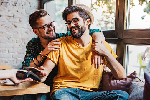 Two gay men pay smiling and using contactless payment in bar