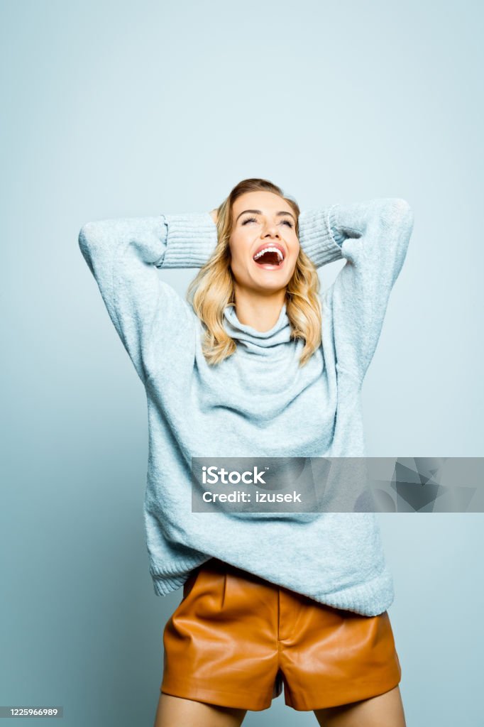Autumn portrait of excited beautiful woman wearing grey sweater Mid adult beautiful woman wearing grey woolen sweater and brown leather shorts standing with raised hands against grey background, looking up and smiling. Women Stock Photo