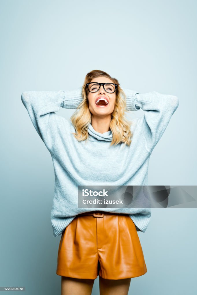 Autumn portrait of excited beautiful woman wearing grey sweater Mid adult beautiful woman wearing grey sweater brown leather shorts and black nerdy glasses standing with raised hands against grey background, looking up and laughing. Women Stock Photo