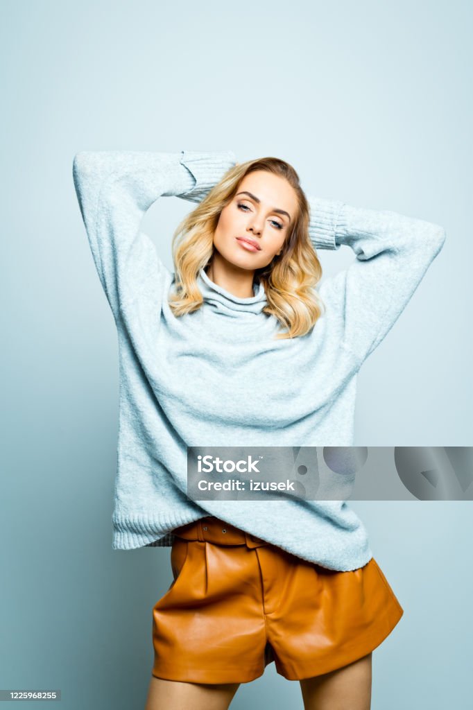 Fashion portrait of beautiful woman wearing grey sweater Autumn portrait of mid adult beautiful woman wearing grey woolen sweater and brown leather shorts standing with raised hands against grey background, looking at camera. 30-34 Years Stock Photo