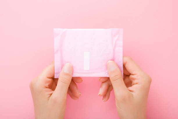 giovane donna mani tenendo confezione di asciugamano sanitario su sfondo tavolo rosa chiaro. colore pastello. primo piano. punto di vista girato. vista dall'alto verso il basso. - sanitary napkin foto e immagini stock