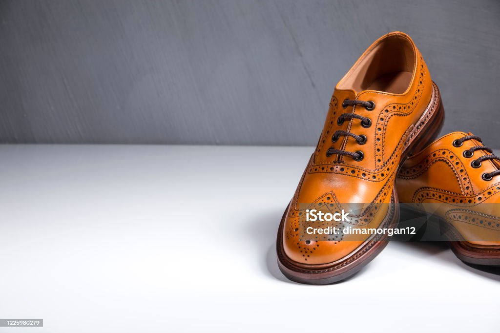Fashionable Luxury Male Full Broggued Tan Leather Oxfords Shoes Placed Over White Surface. Against Gray Wall. Partial View of One Shoe. Horizontal Image Brogue Stock Photo
