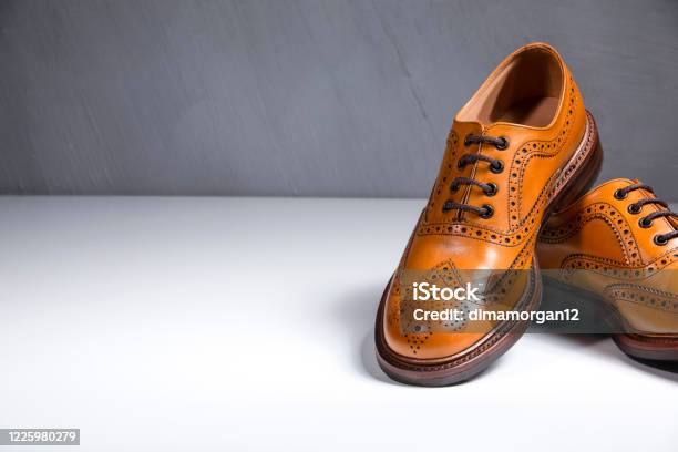 Fashionable Luxury Male Full Broggued Tan Leather Oxfords Shoes Placed Over White Surface Against Gray Wall Partial View Of One Shoe Horizontal Image Stock Photo - Download Image Now