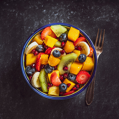 Healthy eating: fresh colorful homemade salad bowl shot from above on dark table. A fork is beside the bowl. Fruits included in the salad are mango, orange, kiwi, strawberry, banana, pomegranate and blueberries. High resolution studio digital capture taken with Sony A7rII and Sony FE 90mm f2.8 macro G OSS lens