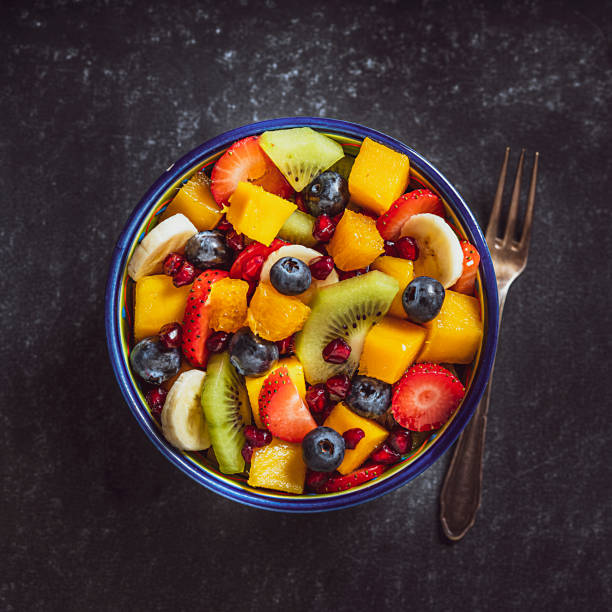 Healthy homemade fruit salad bowl shot from above. Healthy eating: fresh colorful homemade salad bowl shot from above on dark table. A fork is beside the bowl. Fruits included in the salad are mango, orange, kiwi, strawberry, banana, pomegranate and blueberries. High resolution studio digital capture taken with Sony A7rII and Sony FE 90mm f2.8 macro G OSS lens mango fruit photos stock pictures, royalty-free photos & images