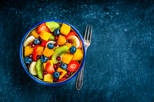 Healthy eating: fresh colorful homemade fruit salad bowl shot from above on dark table. A fork is beside the bowl. Fruits included in the salad are mango, orange, kiwi, strawberry, banana, pomegranate and blueberries. The composition is at the left of an horizontal frame leaving useful copy space for text and/or logo at the right. High resolution 42Mp studio digital capture taken with Sony A7rII and Sony FE 90mm f2.8 macro G OSS lens