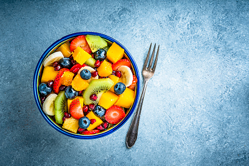 Healthy eating: fresh colorful homemade fruit salad bowl shot from above on bluish tint table. A fork is beside the bowl. Fruits included in the salad are mango, orange, kiwi, strawberry, banana, pomegranate and blueberries. The composition is at the left of an horizontal frame leaving useful copy space for text and/or logo at the right. High resolution 42Mp studio digital capture taken with Sony A7rII and Sony FE 90mm f2.8 macro G OSS lens
