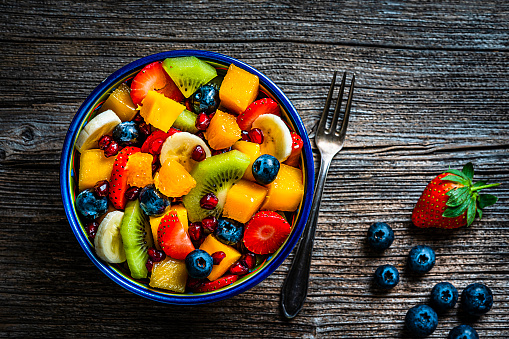 Healthy eating: fresh colorful homemade salad bowl shot from above on rustic wooden table. A fork is beside the bowl. Fruits included in the salad are mango, orange, kiwi, strawberry, banana, pomegranate and blueberries. The composition is at the left of an horizontal frame leaving useful copy space for text and/or logo at the right. High resolution 42Mp studio digital capture taken with Sony A7rII and Sony FE 90mm f2.8 macro G OSS lens
