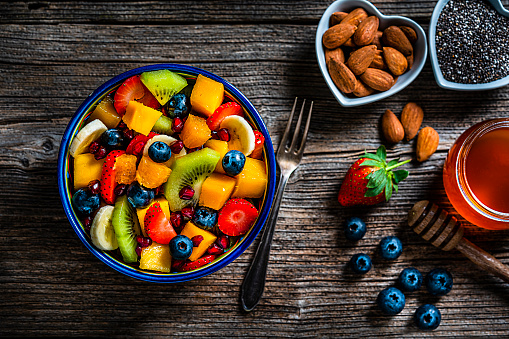 Healthy eating: fresh colorful homemade fruit salad bowl shot from above on rustic wooden table. Two heart shaped bowls with chia seeds and almonds and a honey jar are beside the fruit salad bowl. A fork complete the composition. Fruits included in the salad are mango, orange, kiwi, strawberry, banana, pomegranate and blueberries.  High resolution 42Mp studio digital capture taken with Sony A7rII and Sony FE 90mm f2.8 macro G OSS lens