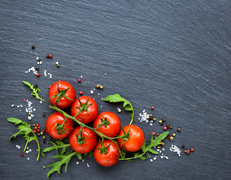 Fresh cherry tomatoes, salt, pepper and arugula leaf on a black slate board, top view