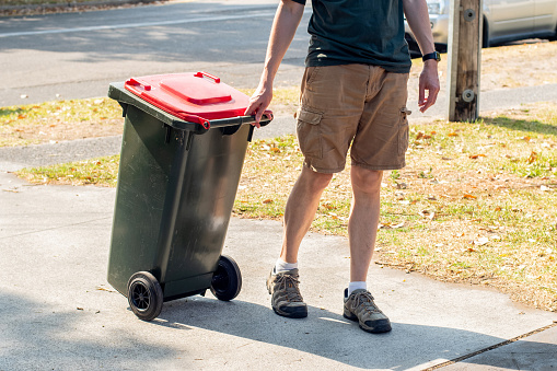 A man dragging the household wheelie red bin with general waste on the street for council gargbage collection. Waste management concept.