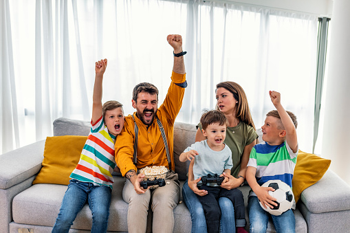 Playful family playing video games together in a living room while holding soccer ball.