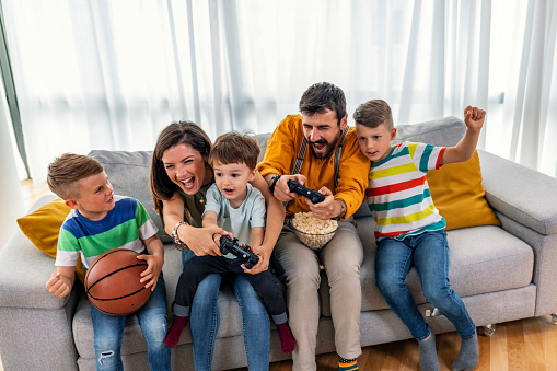 Laughing family playing video games in a living room. Happy family using joysticks and having fun while holding basketball ball.