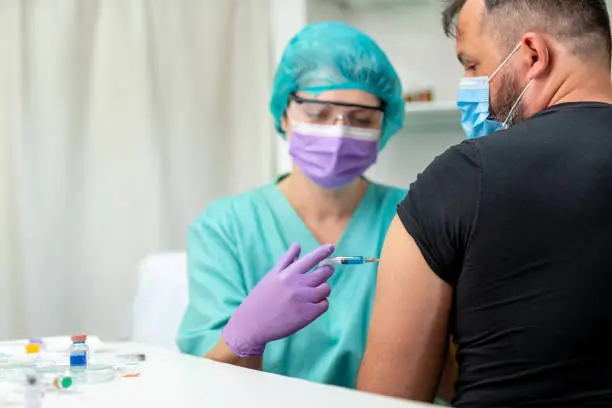 Photo of Mid adult man getting flu shot, nurse in protective workwear injecting vaccine