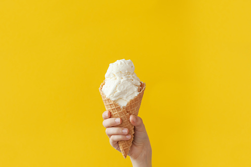 Children's hand holds delicious vanilla ice cream cone on a bright yellow background with copy space.